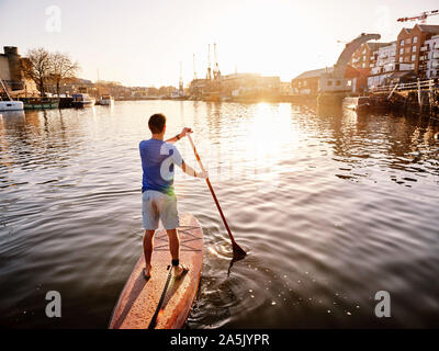 Uomo in piedi su paddleboard sul fiume all'alba, inquadratura da dietro Foto Stock