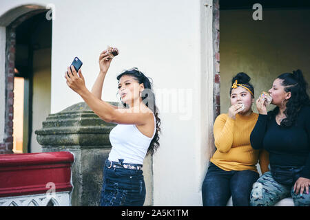 Giovane donna prendendo selfie con sorelle adolescenti mentre mangia torte in porta Foto Stock