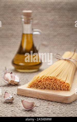 Olio di cottura e ingredienti. Spaghetti legati in mazzi da corda artigianale giacente sulla superficie naturale bulbi di aglio. Caraffa con olio di oliva sullo sfondo Foto Stock