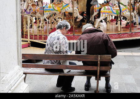 Elderely matura in appoggio sul banco di fronte a merry go round su una tranquilla aternoon in una città europea Foto Stock