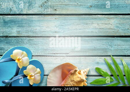 Piatto estivo sullo sfondo dei laici. Tropical foglie di palma, flip flop e fiore sul vecchio blu sullo sfondo di legno. Foto Stock