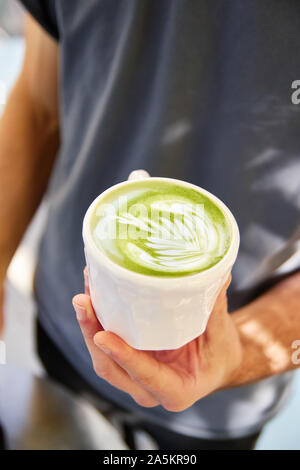 Voce maschile barista proponendo piatti vegan matcha cappuccino con Latte di avena con latte art in outdoor cafe. Foto Stock