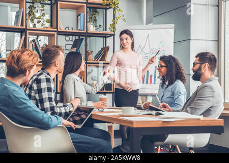 Startupers lavorando insieme in ufficio seduti a discutere il progetto allegro mentre la donna incinta in piedi spiegare i dati Foto Stock