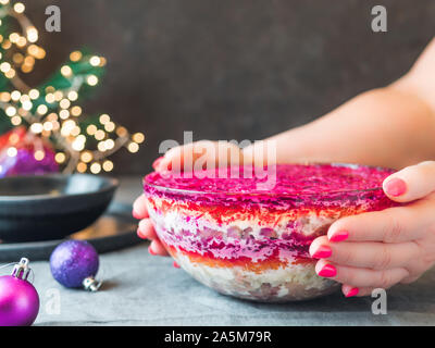 Donna mani con insalata stratificata aringa sotto una pelliccia sulla tavola festiva. Tradizionale insalata russa con le aringhe e verdure in recipiente di vetro. Copia spazio per il testo. Foto Stock