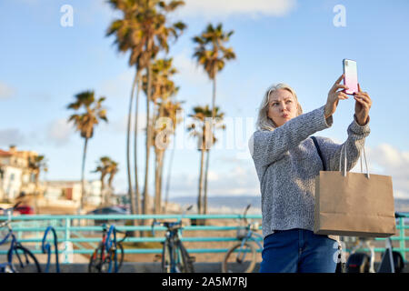 Senior donna raggrinzamenti labbra tenendo selfie con smart phone contro sky durante il tramonto Foto Stock