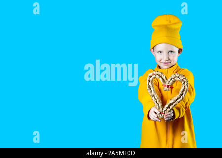 Un piccolo bambino carino in una giacca gialla e cappello. Tenendo un cuore intrecciato su un brillante sfondo colorato. Foto Stock