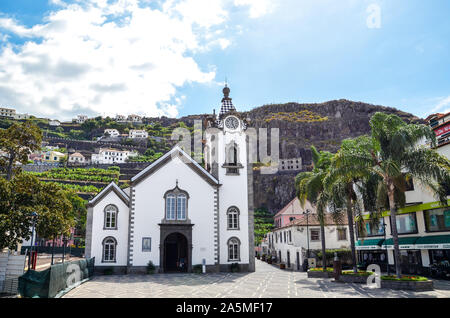 Ribeira Brava, Madeira, Portogallo - 9 Sep, 2019: bella chiesa cattolica romana nel centro della citta'. Palme sulla piazza. Piantagioni di banane su una collina rocciosa in background. Foto Stock