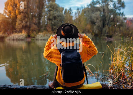 Viaggiatore con zaino rilassante da autunno river al tramonto. Giovane donna seduta sul banco e rilassante azienda hat. Uno stile di vita attivo Foto Stock