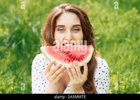 Bella ragazza è mangiare pezzo di rosso anguria maturi nella natura dello sfondo. Giovane donna mangia pezzo di anguria su sfondo verde d'erba. Foto Stock