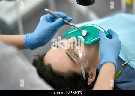 Ragazza carina in occhiali protettivi e una guancia avvolgitore con una gomma verde di diga in una clinica dentale. Dentista di guanti in lattice blu esamina i suoi denti con Foto Stock