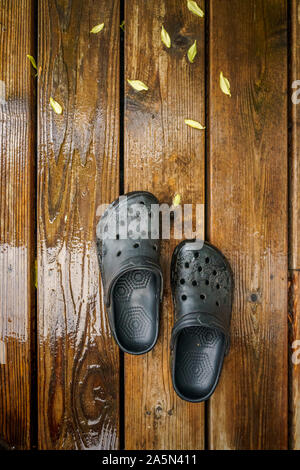 POV di due nero in gomma plastica ostruiscono il giardinaggio sandali sulla parte superiore di legno umido decking con giallo Foglie di autunno. Foto Stock