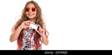 Teen ragazza con lunghi capelli biondi tinti con suggerimenti rosa, in shiny abito leggero, nero e bianco sneakers, bicchieri, in piedi con le cuffie, tenendo in mano una carta Foto Stock