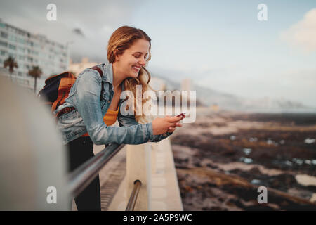 Una bionda sorridente giovani viaggiatori femmina con il suo zaino appoggiato sulla ringhiera in via della città utilizzando il suo telefono cellulare Foto Stock