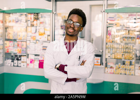 Bel giovane americano africano uomo farmacista in bicchieri e mantello bianco in piedi nella parte interna della farmacia moderna Foto Stock