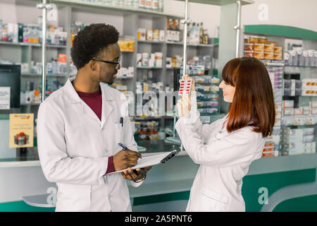 L'uomo africano il farmacista iscritto sulla clipboard mentre il suo collega femmina trattiene un blister di pillole di rosso nelle mani, il controllo della qualità dei medicinali Foto Stock