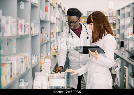 Ritratto di due sorridenti multiethnical amichevole ai farmacisti che lavorano in farmacia moderna e facendo ordine per medicinali in compagnia di distribuzione Foto Stock