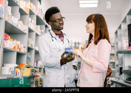 Bel maschio africano il farmacista parlando con una bellissima femmina client in luce moderno farmacia. Foto Stock