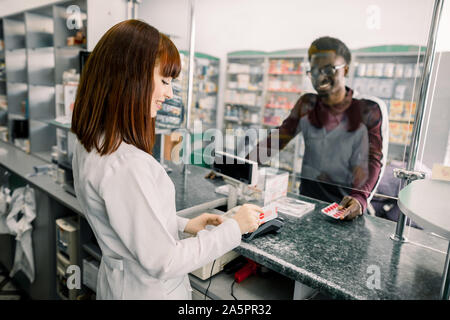 Giovane africano di pagare per pillole con carta di credito in farmacia. Donna sorridente al farmacista dando pillole di uomo nero in farmacia Foto Stock