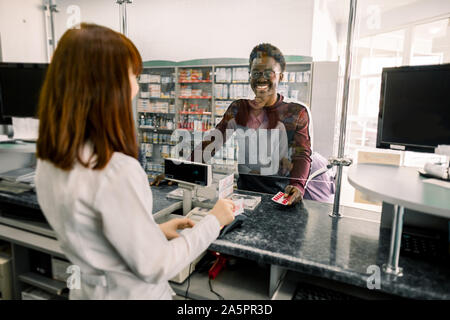 Giovane africano di pagare per pillole con carta di credito in farmacia. Donna sorridente al farmacista dando pillole di uomo nero in farmacia Foto Stock