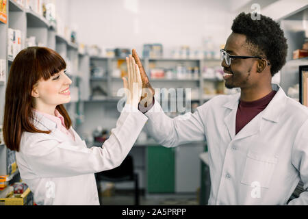 Due lieti colleages farmacisti africana di uomo e donna caucasica lavora in farmacia, sorridente. dando cinque e divertirsi. Foto Stock