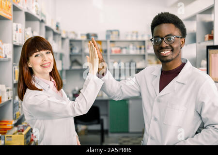 Due lieti colleages farmacisti africana di uomo e donna caucasica lavora in farmacia, sorridente. dando cinque e divertirsi. Foto Stock