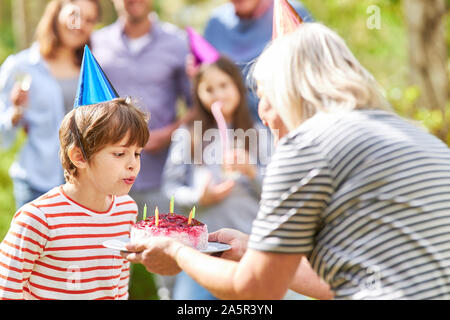 Il ragazzo si brucia le candele sulla sua torta di compleanno in giardino in estate Foto Stock