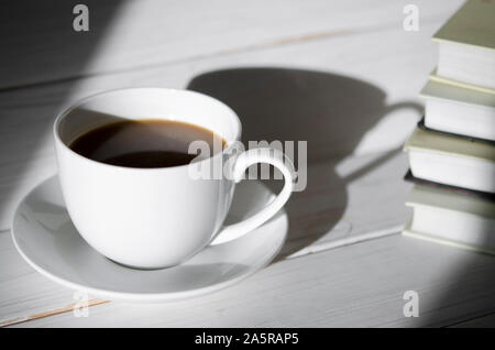 Un bianco rotondo tazza di caffè si erge su un piattino bianco su bianco di un tavolo di legno accanto all'ombra della tazza Foto Stock