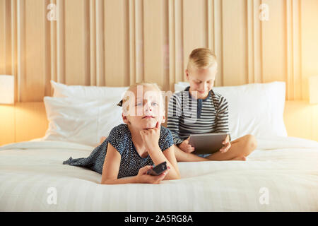 Incantevole piccola ragazza distesa su un letto a guardare la televisione con il suo fratello seduto dietro di lei utilizzando una tavoletta digitale Foto Stock