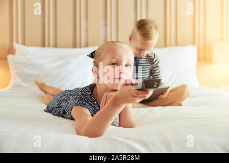 Incantevole piccola ragazza distesa su un letto cambiando i canali mentre si guarda la televisione con suo fratello seduto dietro di lei utilizzando una tavoletta digitale Foto Stock