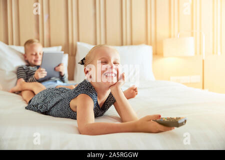 Ridere bambina giacente su un letto a guardare la televisione con il suo fratello seduto dietro di lei utilizzando una tavoletta digitale Foto Stock