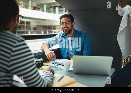 Ridendo gruppo di diversi progettisti che lavorano insieme su un progetto in una riunione pod nella lobby di un ufficio moderno Foto Stock