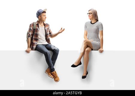 A piena lunghezza shot di un adolescente studente maschio parlando a un youn g donna seduta su un pannello isolato su sfondo bianco Foto Stock