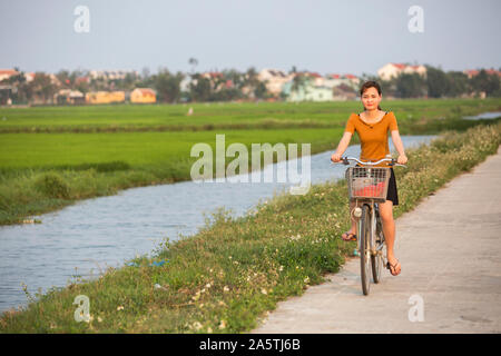 Una giovane donna corse una bicicletta passato un risone in Hoi An, Vietnam. Foto Stock