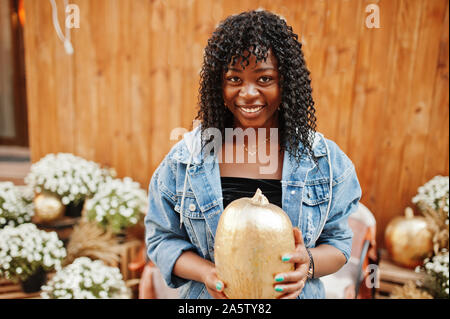 Alla moda ed elegante afro Francia curly donna poste in autunno il giorno in jeans giacca. Africano nero modello femminile con la zucca a mani. Foto Stock