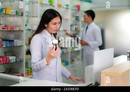 Fiducioso asiatici femmina giovane farmacista con sorriso tenendo una bottiglia di medicina e ricerca di quel prodotto nel database di computer in farmacia farmaci Foto Stock