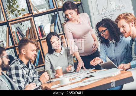 Startupers lavorando insieme a Office seduta guardando i documenti felice mentre la donna incinta in piedi per spiegare l'analisi dei dati Foto Stock