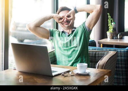 Giovani funny crazy businessman in verde t-shirt seduto e guardando con il binocolo gesto. business e concetto freelancing. interna shot vicino grande vittoria Foto Stock