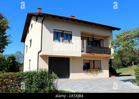 Rinnovato di recente famiglia suburbana a casa con balcone anteriore e piastrelle di pietra carraio circondato con siepe e alberi sul cielo azzurro sfondo Foto Stock