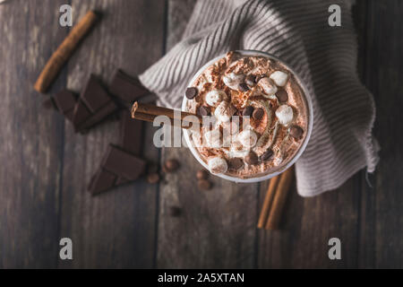 Un boccale di bianco di lusso con cioccolata calda con panna montata e pezzi di marshmallows e scaglie di cioccolato. Bastoncini di cannella e ieces del cioccolato sono Foto Stock