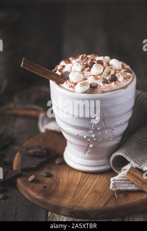 Un boccale di bianco di lusso con cioccolata calda con panna montata e pezzi di marshmallows e scaglie di cioccolato. Pezzi di cioccolato sono sparsi intorno o Foto Stock