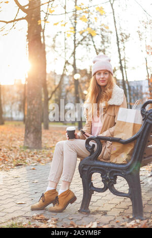 Redhead girl in vestiti caldi seduta con caffè sul banco di legno in autunno park. Foto Stock