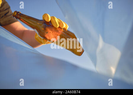 La mano di una donna in un giallo Guanto in gomma mette un usato bottiglia in vetro marrone in un cestino blu borsa. Blue sky. Persone e di ecologia. Riverside inquinamento. Preser Foto Stock