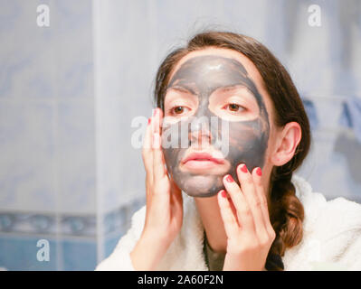 Cosmetici maschera nera. La donna porta la bellezza per il suo volto si sbarazza di puntini neri, utilizzando una maschera nera. Una femmina in bagno guarda nello specchio Foto Stock