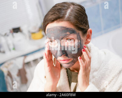 Cosmetici maschera nera. La donna porta la bellezza per il suo volto si sbarazza di puntini neri, utilizzando una maschera nera. Una femmina in bagno guarda nello specchio Foto Stock