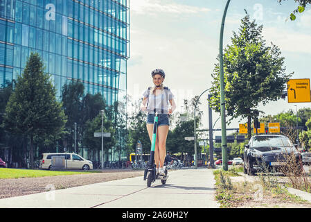 Giovane donna in sella E-scooter sul marciapiede, Berlino, Germania Foto Stock