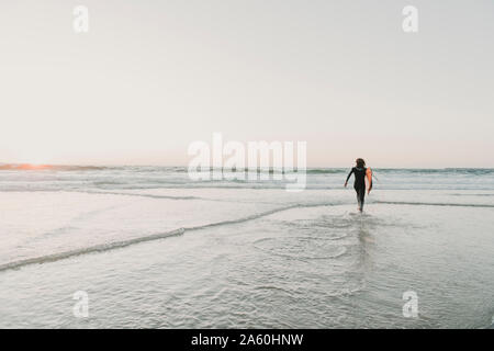 Vista posteriore del surfista in esecuzione sulla spiaggia al tramonto, Costa Nova, Portogallo Foto Stock