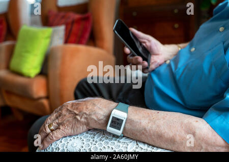 Uomo anziano utilizzando un telefono cellulare e che indossa una smart allarme di emergenza bracciale da polso intorno a casa Foto Stock