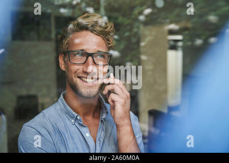 Sorridente giovane imprenditore al telefono dietro il vetro di finestra di office Foto Stock