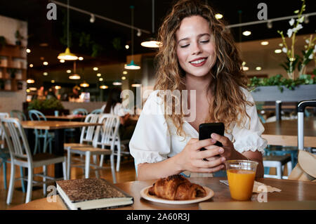 Sorridente giovane donna utilizza lo smartphone in una caffetteria pur avendo prima colazione Foto Stock