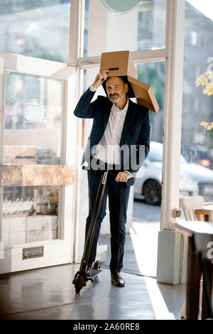 Imprenditore con scatola di cartone sulla sua testa, equitazione e-scooter in un coffee shop Foto Stock